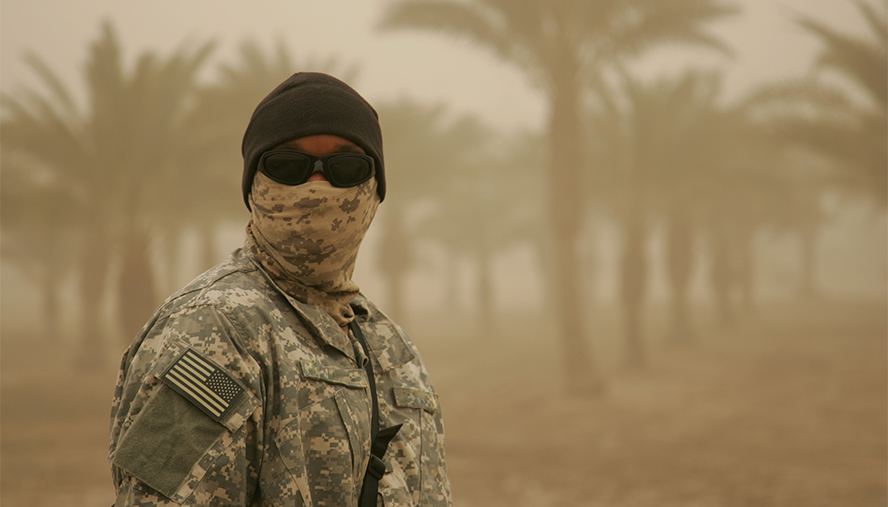 Man in camoflage standing in a sandy dessert with palm trees